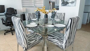 A modern dining area with a glass table surrounded by zebra-striped chairs, a vase with yellow flowers, and a black office chair in the background.