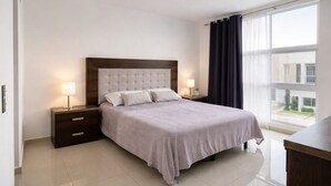 Quarto de hotel moderno com uma cama grande, mesa de cabeceira com abajur, cômoda e vista para o exterior através de uma janela com cortinas escuras.