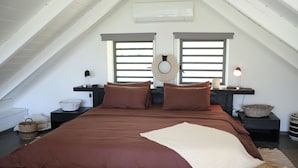 Une chambre à coucher avec un grand lit, deux tables de chevet, un climatiseur fixé au mur et une fenêtre avec des stores.
