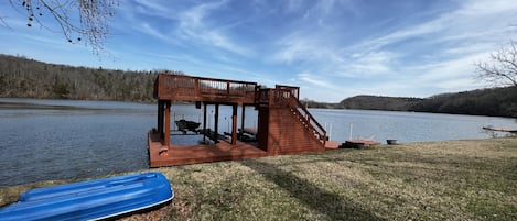 A wooden deck with a staircase leading to a covered area by a lake, a boat docked nearby, and a blue kayak on the grass.