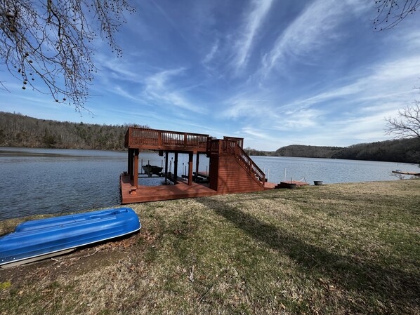 A wooden deck with a staircase leading to a covered area by a lake, a boat docked nearby, and a blue kayak on the grass.