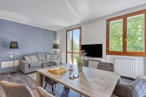 Ein modernes Wohnzimmer mit Sofa, Esstisch, Fernseher und großen Fenstern.
