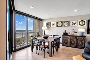Un espace repas comprenant une grande table et des chaises, une vue sur l’océan grâce à de larges fenêtres, et un meuble décoratif où sont exposées des œuvres d’art.