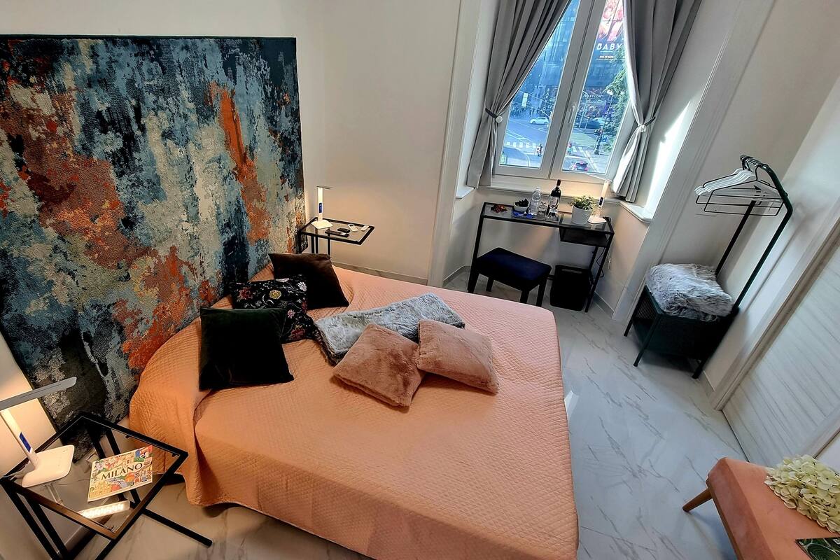 Une chambre avec un grand tableau abstrait, un lit avec de nombreux coussins, un bureau avec une chaise et une fenêtre avec des rideaux.