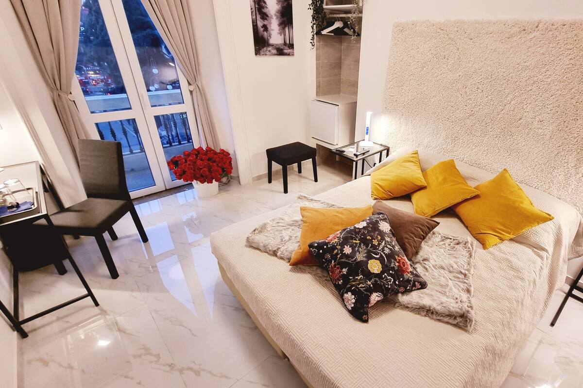 Une chambre moderne avec un grand lit, un bureau et une chaise. Une fenêtre offre une vue sur le paysage urbain.