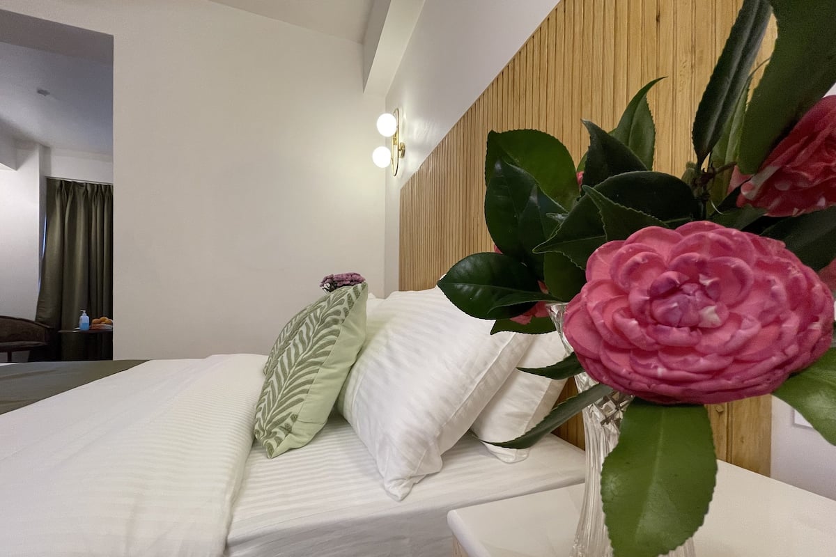 Una habitación de hotel con una cama, una mesita de noche, un jarrón con una flor y un cabecero de madera.