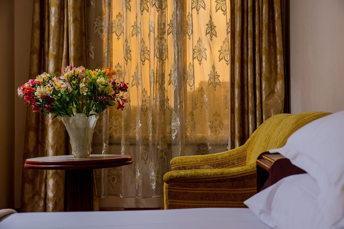 Habitación de hotel con cama, una mesita redonda con un florero y una silla con cojín estampado.