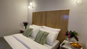 Una cama bien tendida con sábanas blancas y almohadas estampadas en verde, una mesita de noche con un florero y una cesta de naranjas, y lámparas fijadas a la pared.