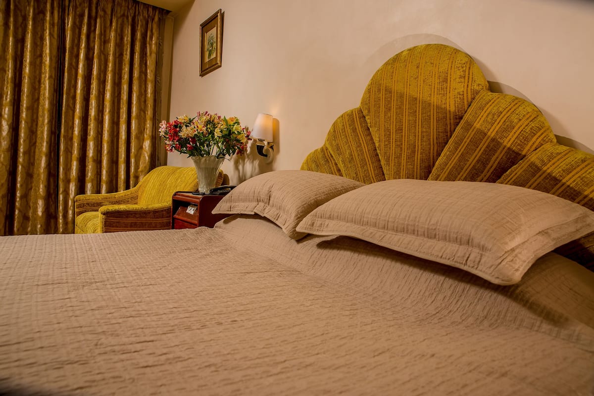 Una cama con ropa de cama color beige, un sillón amarillo, una mesita de noche con una lámpara y un jarrón con flores.