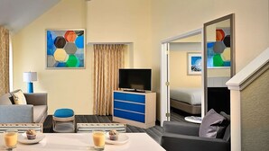 Une chambre d’hôtel avec un lit, une télévision, un ventilateur de plafond, une table basse avec deux muffins et un verre de café, un canapé, une armoire bleue et un tableau accroché au mur.