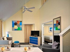 Quarto de hotel com cama, televisão, ventoinha no teto, mesa de centro com dois muffins e uma chávena de café, sofá, armário azul e um quadro na parede.