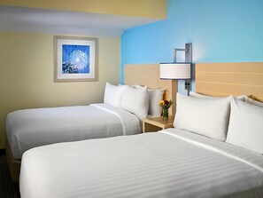Quarto de hotel com duas camas, uma mesa de cabeceira com abajur, um quadro na parede e um vaso com flores.