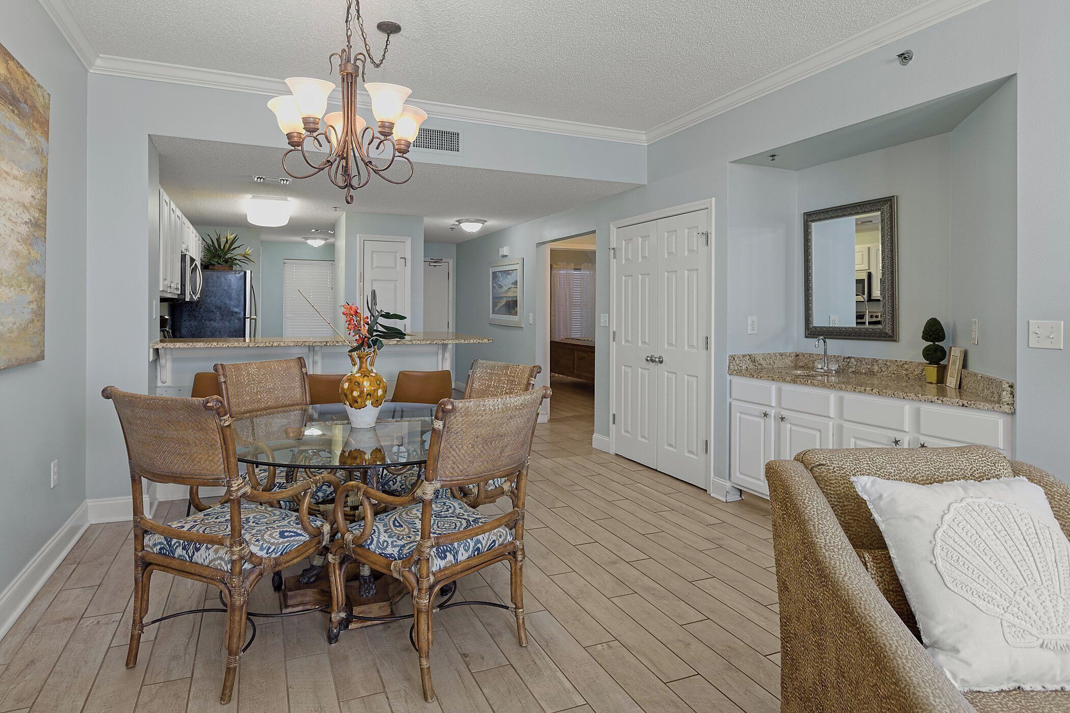 A dining area with a glass table and wicker chairs, a kitchen with white cabinets, and a living room with a beige sofa.