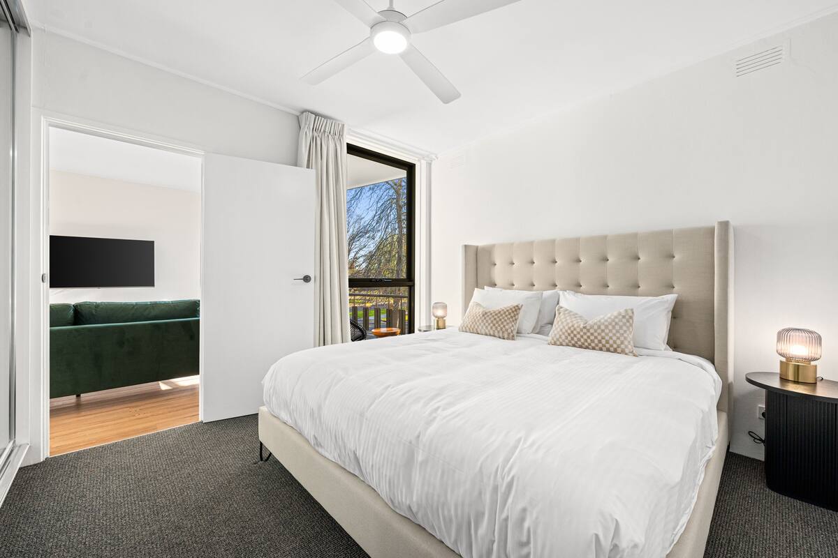 Quarto com uma cama grande, um ventilador de teto, uma mesa de cabeceira com abajur e vista para o exterior através de uma porta aberta.