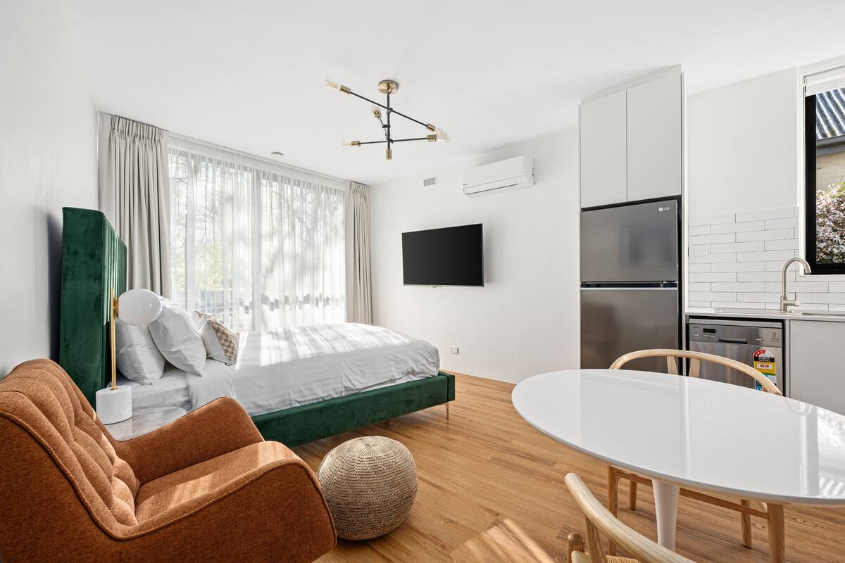 Quarto moderno com cama, parede de destaque verde, TV e área de cozinha com frigorífico e máquina de lavar louça.