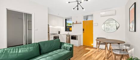 Un apartamento moderno con un sofá verde, una mesa de comedor con sillas, una zona de cocina y una puerta amarillo brillante.