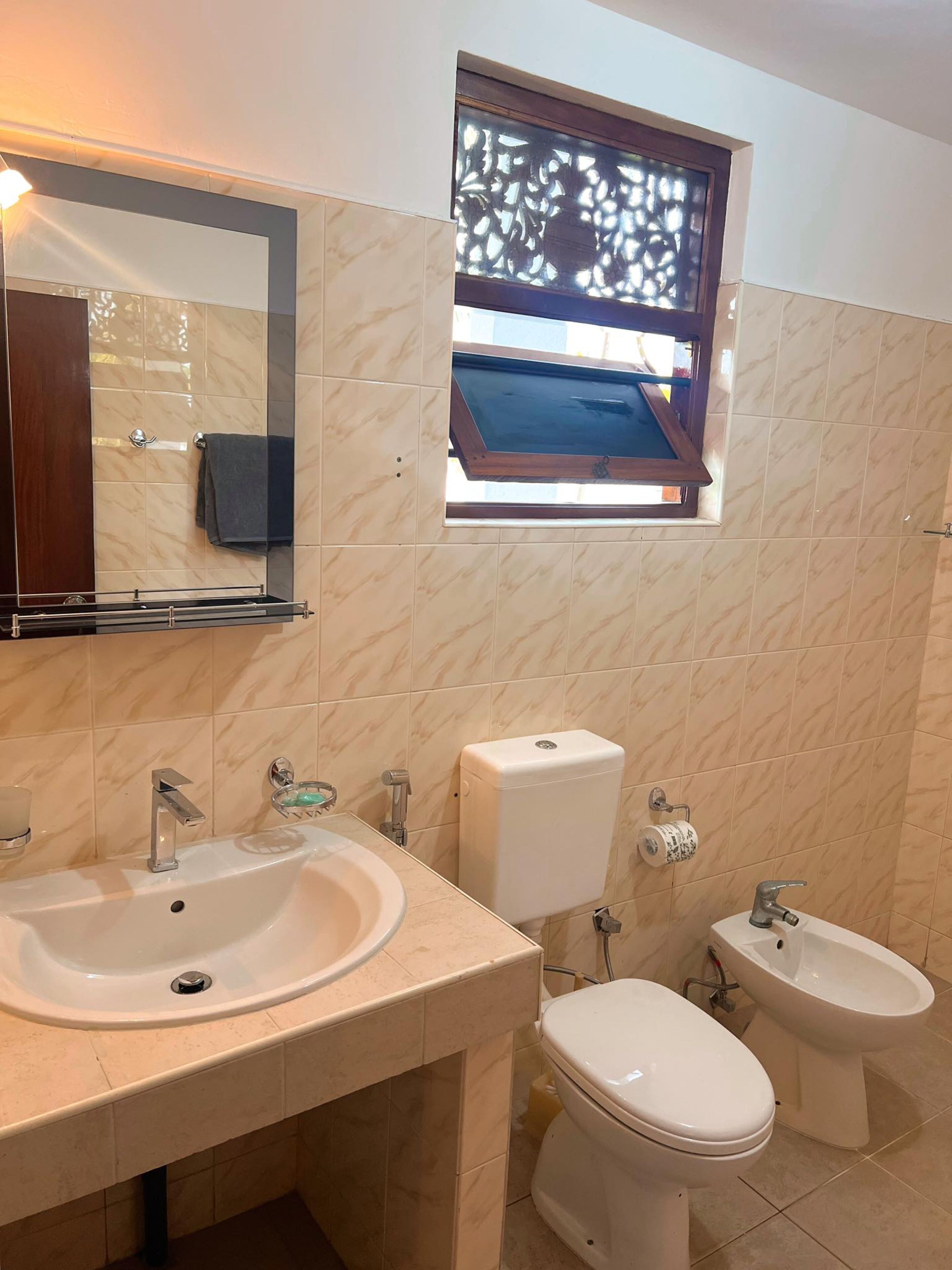 A bathroom with a sink, toilet, bidet, and a window with a decorative screen.