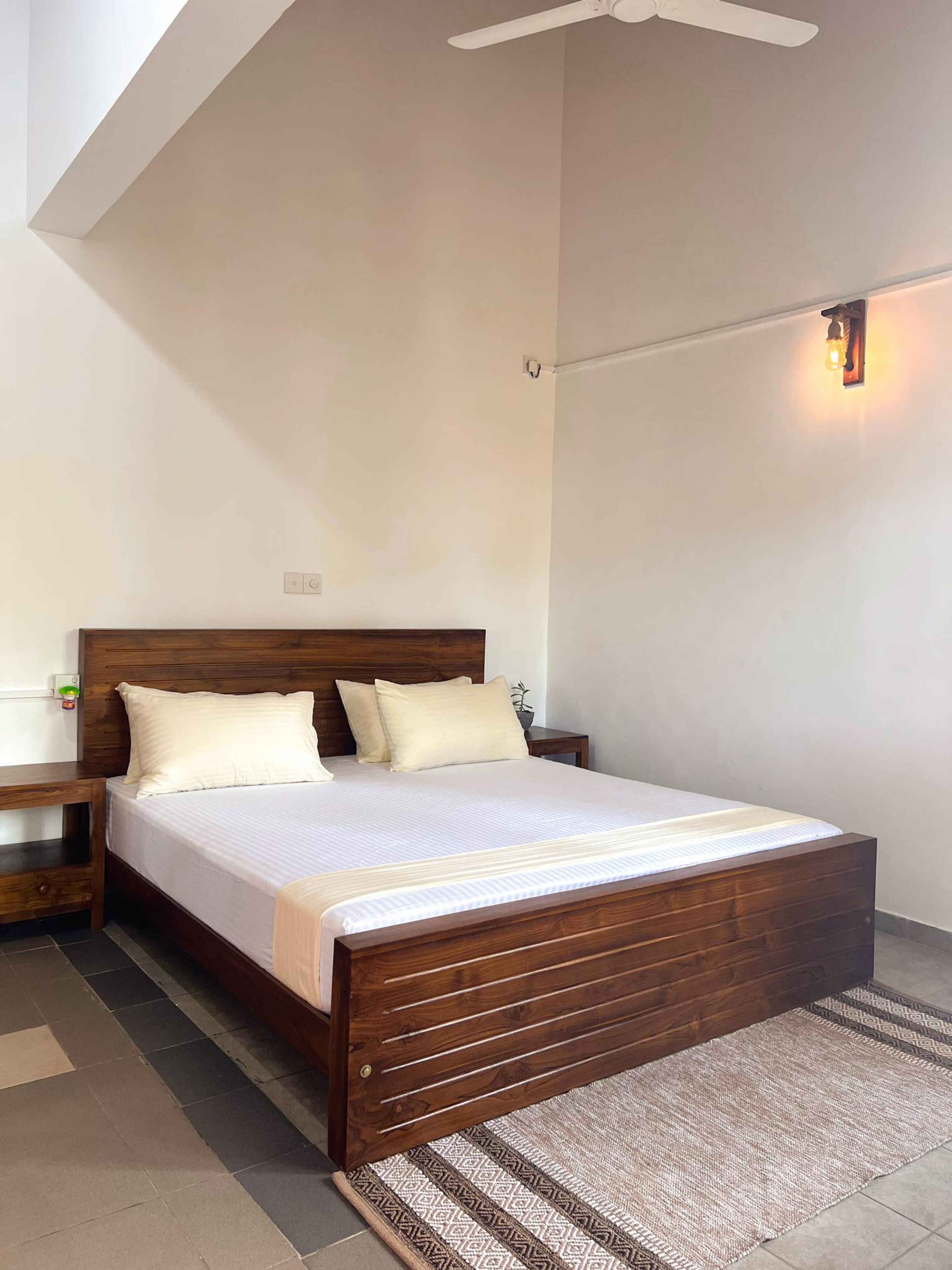 Un dormitorio con una cama de madera, ropa de cama blanca, una mesita de noche, una lámpara fijada a la pared y un ventilador de techo.
