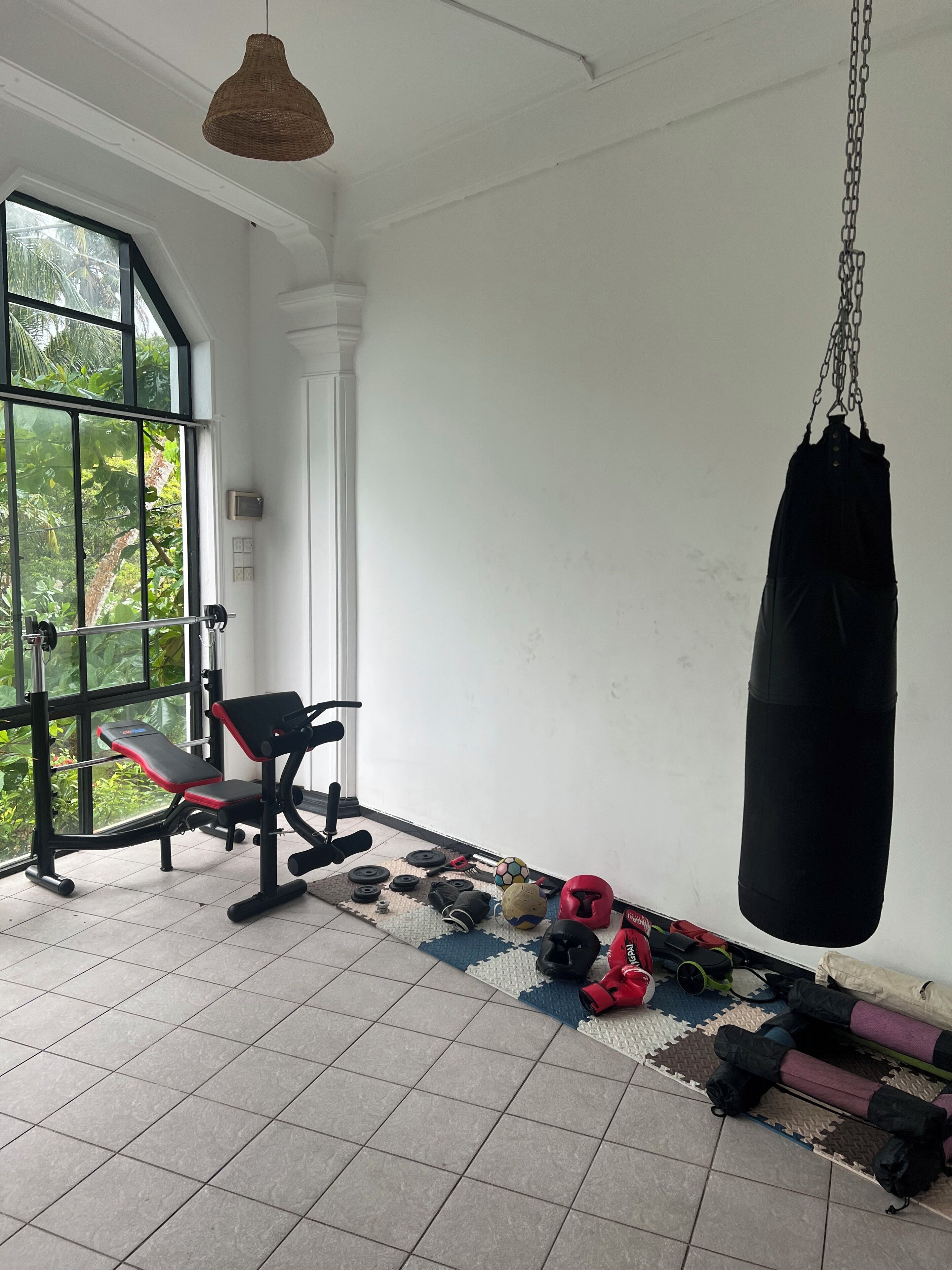 A gym with a bench, weights, a punching bag, and a window with a view of greenery.
