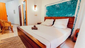 A bedroom with a wooden bed, white bedding, a headboard with a blue abstract painting, a wooden nightstand, and a wicker chair. - Hummus Hotel Hikkaduwa  (Hikkaduwa)