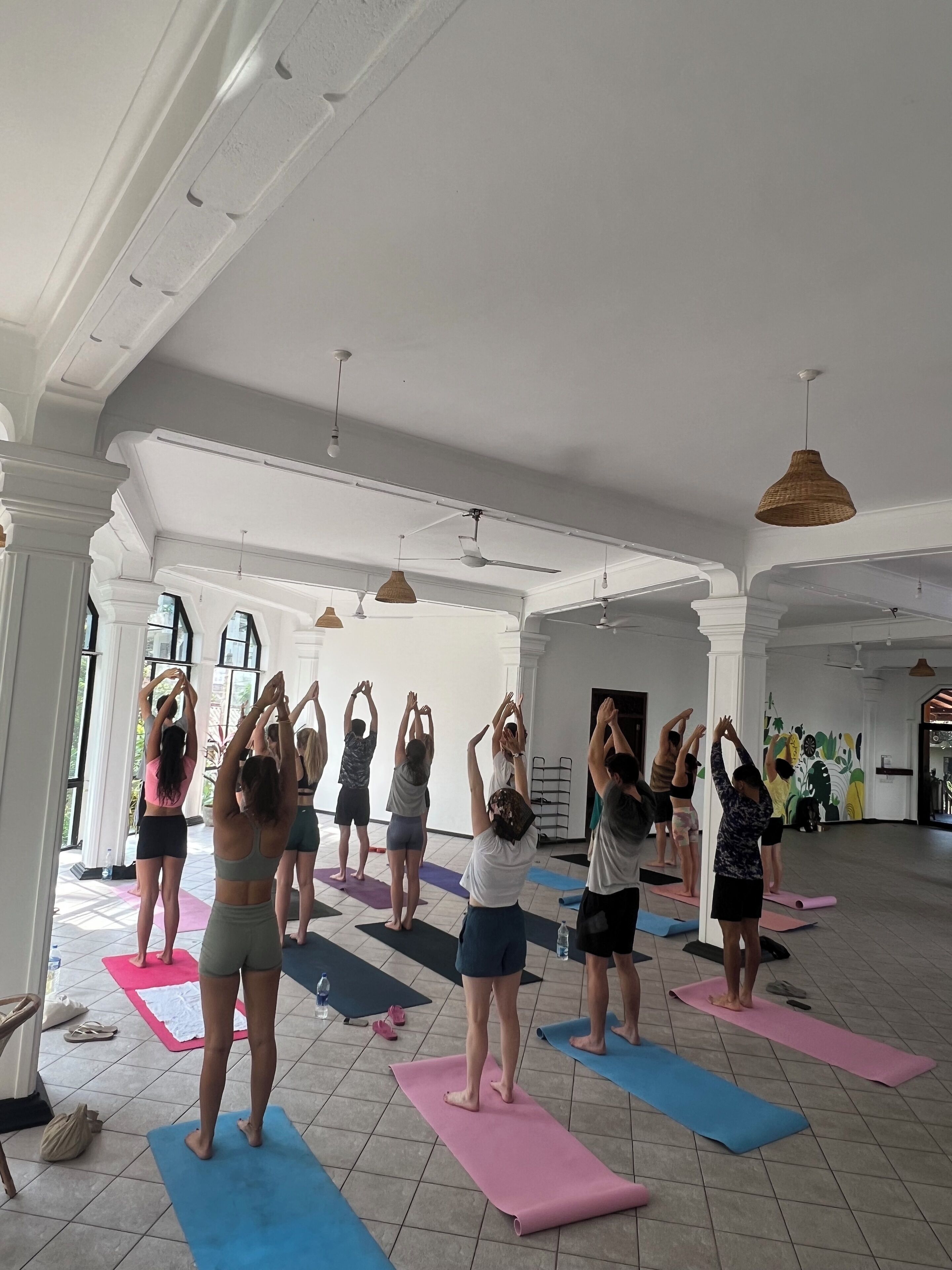 A spacious room with high ceilings and large windows, featuring a group of people on yoga mats performing a synchronized stretch.
