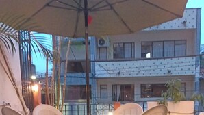 Eine Terrasse mit Tisch und Stühlen unter einem großen Sonnenschirm, umgeben von Blumentöpfen und einem Gebäude im Hintergrund.