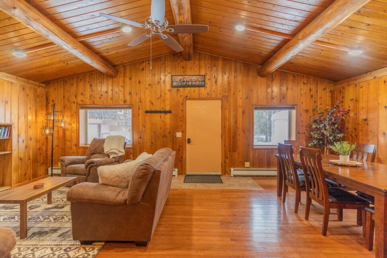 Ein gemütliches Blockhaus-Interieur mit Holzbalkendecke, Deckenventilator, Kamin, Esstisch mit Stühlen und einer Couch.