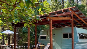 A rustic outdoor seating area with wooden chairs and a picnic table, a covered wooden structure, and a vintage-style RV. - Radio Ranch (Burnt Ranch)