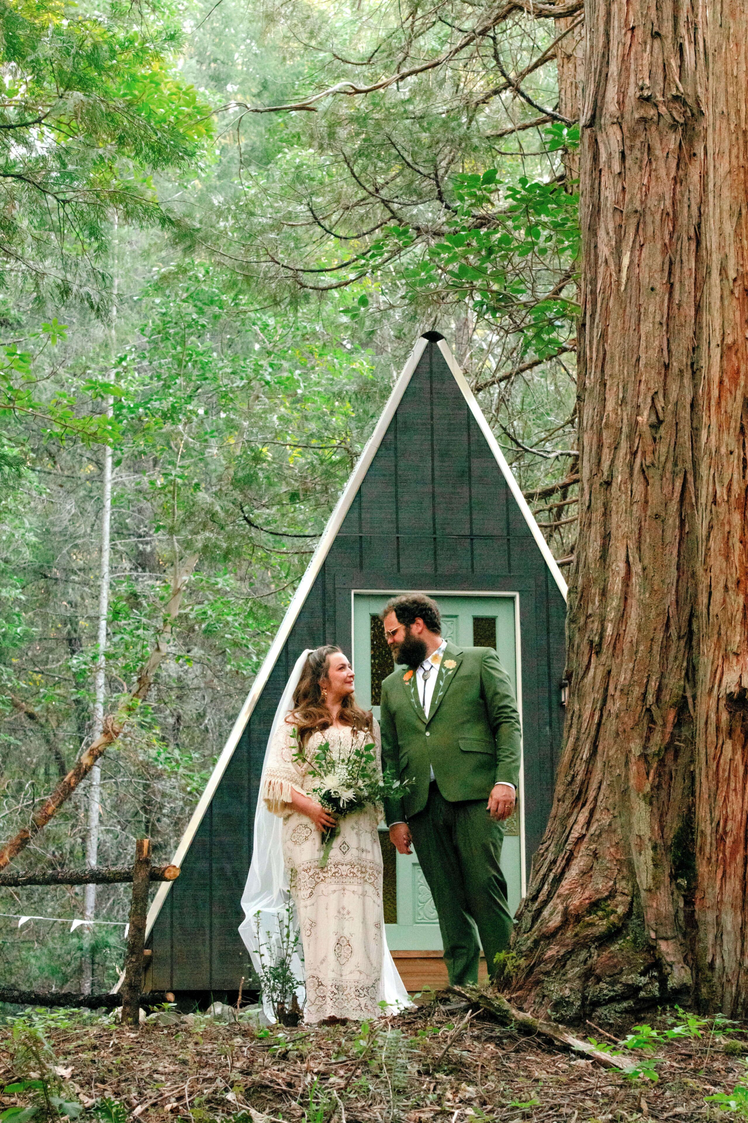 A wedding couple in front of a small wooden cabin in a forest.