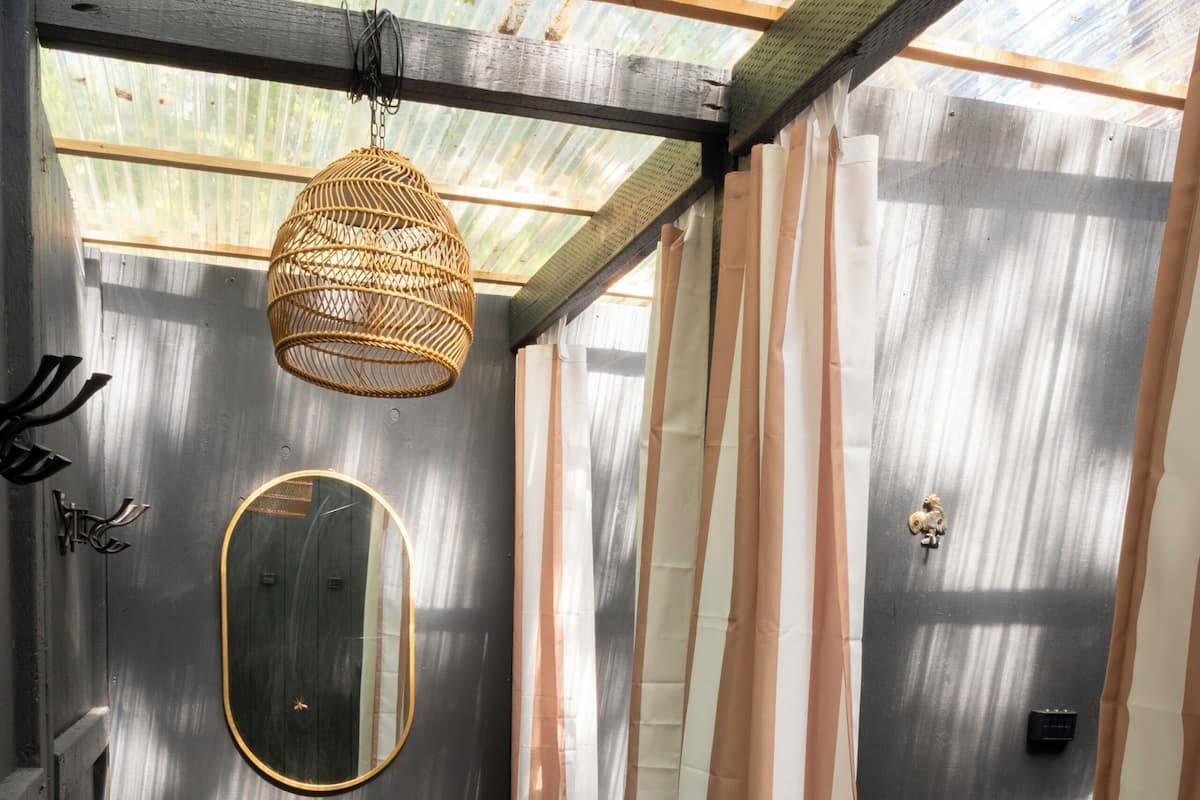 A small, enclosed space with a metal-framed ceiling, a wicker hanging light fixture, a mirror, striped curtains, a potted plant, and a basket labeled "TOWELS".