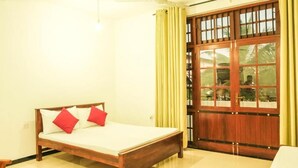 A room with a wooden bed, yellow curtains, and a large wooden door with glass panels. - Cottage 23 (Kurunegala)