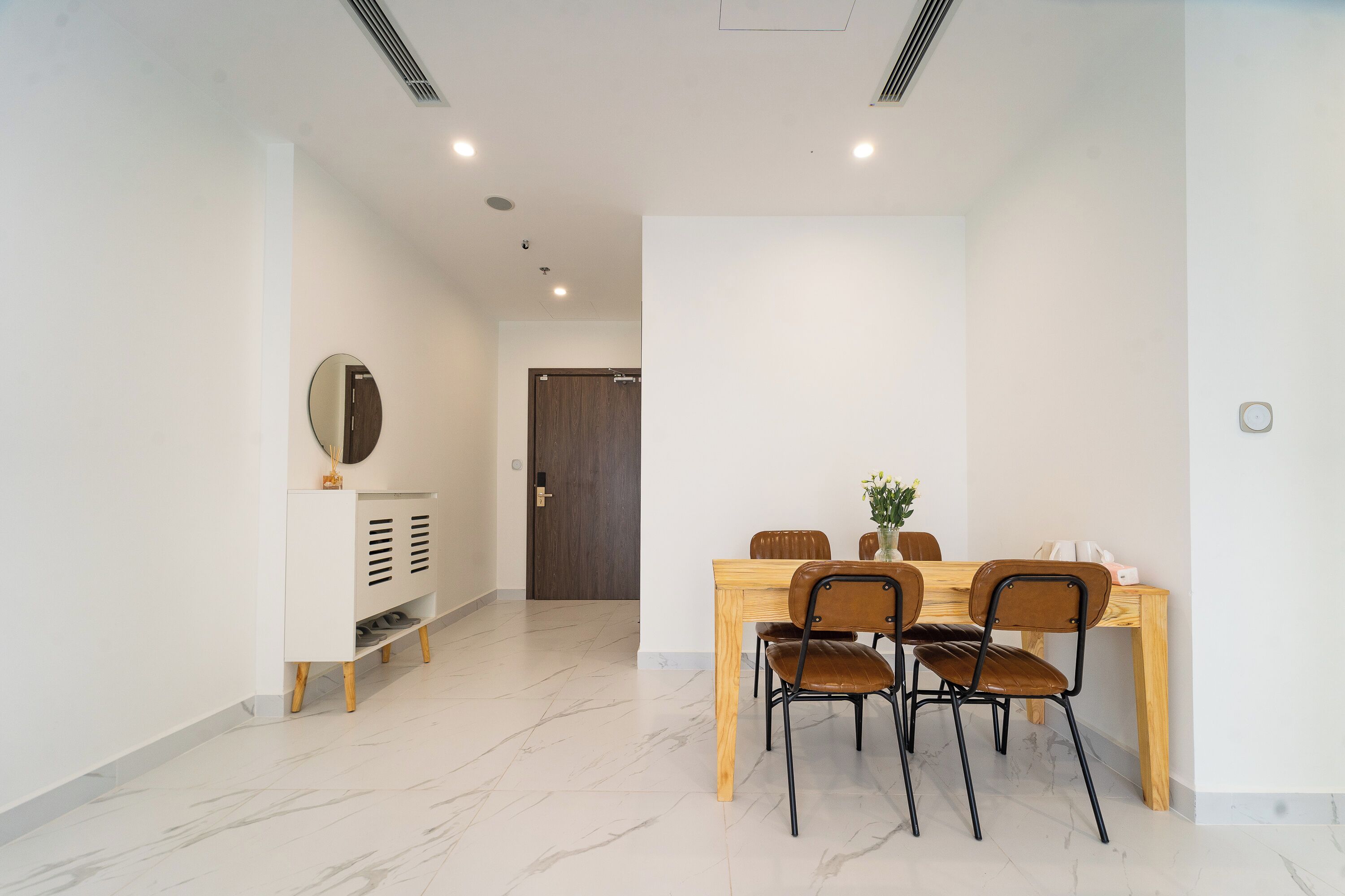 A dining area with a wooden table and four chairs, a white console with a mirror, and a door in the background.
