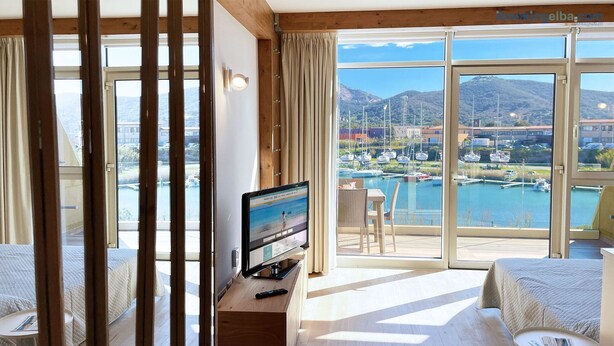 A hotel room with a large window offering a marina view, a television on a wooden table, and two beds with white bedding. - Fronte Mare Suites - Fronte Mare Suites - Studio (H) (Portoferraio)