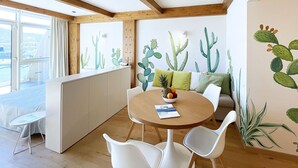 A modern room with a wooden ceiling, a round dining table with chairs, a sofa with cushions, and a wall with cactus illustrations. - Fronte Mare Suites - Fronte Mare Suites - Studio (H) (Portoferraio)