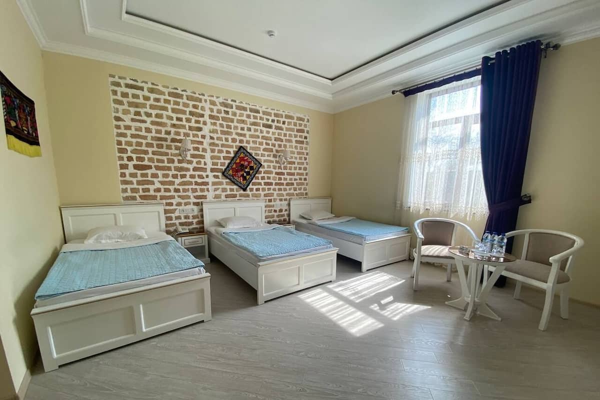 A room with three single beds, a small table with chairs, a brick-patterned wall, and a window with curtains.