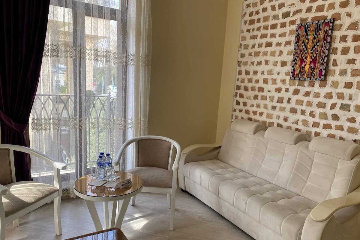 A living room with a white sofa, two white armchairs, a glass coffee table with water bottles, and a patterned artwork on a brick wall.