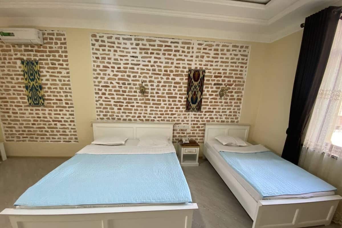 A room with two beds, a brick-patterned wall, a small bedside table, and a window with curtains.