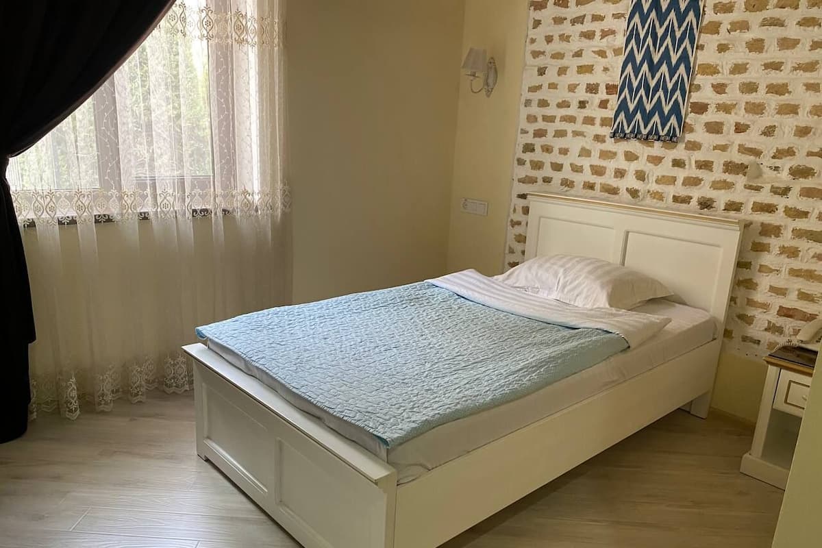 A single-bed room with a white headboard, a patterned bedspread, a wooden floor, a window with sheer curtains, and a brick-patterned wall.