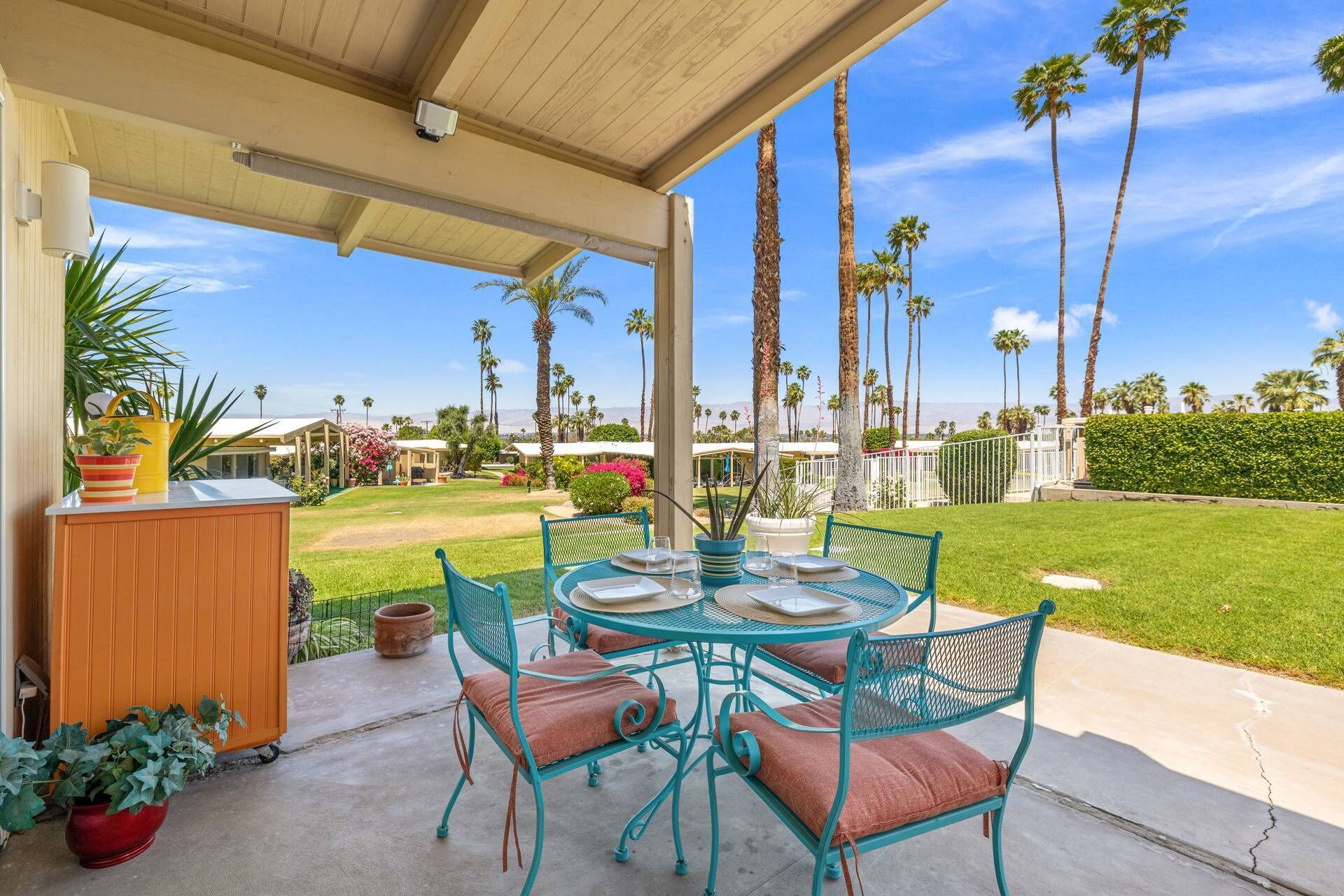 A covered patio with a round table and chairs, surrounded by potted plants and a view of palm trees and houses.