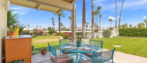A covered patio with a round table and chairs, surrounded by potted plants and a view of palm trees and houses.