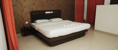 A neatly made bed in a hotel room with a dark headboard, red curtains, and a patterned wallpaper.