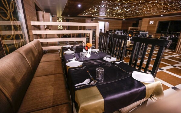 A restaurant with a long table set for dining, cushioned seating, and a decorative ceiling. - Hotel Trupti International Hubli (Hubli)