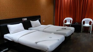 Een hotelkamer met twee enkele bedden, een houten hoofdbord, een rood gordijn en twee witte plastic stoelen.