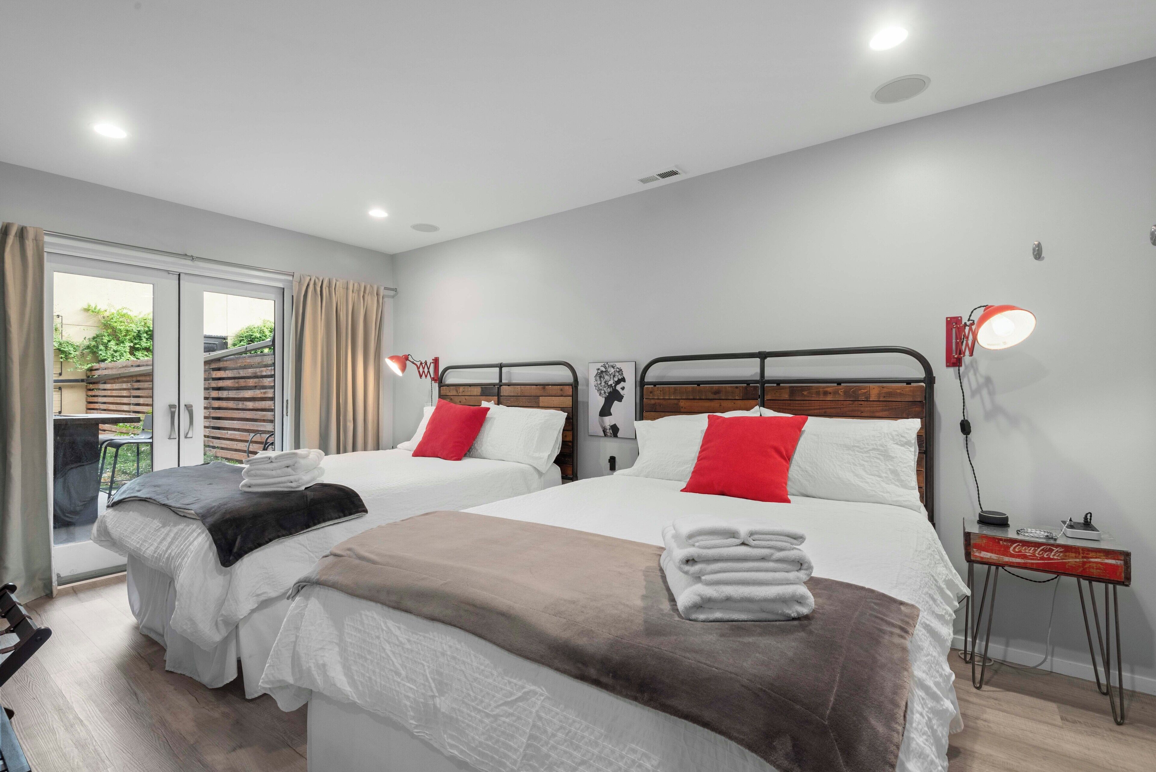 A modern hotel room with two beds, a sliding glass door to the outside, a small table with a lamp, and a wall-mounted artwork.