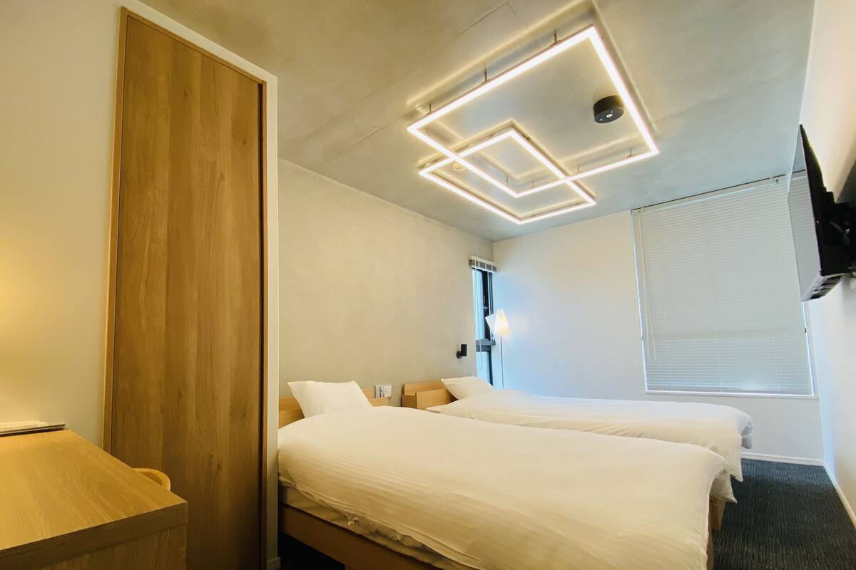 A modern hotel room with two beds, a wooden wardrobe, a desk, and a flat-screen TV mounted on the wall.