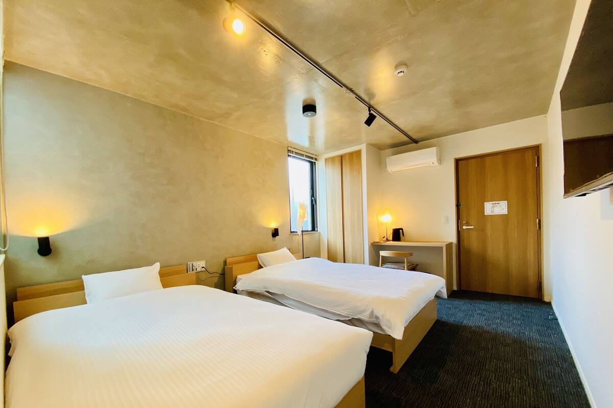 A hotel room with two beds, a desk, a chair, and a door.