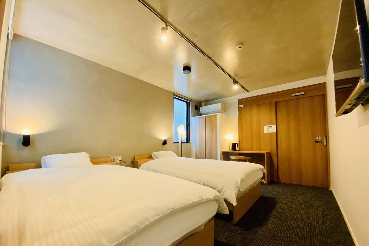 A hotel room with two beds, a desk, a chair, a wardrobe, and a window.