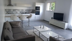 Un salon moderne avec un canapé, une table basse et un téléviseur. La cuisine est équipée d’un four à micro-ondes, d’un four et d’un plan de travail carrelé.