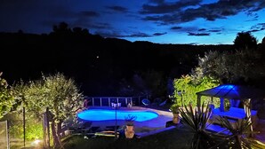 Une vue nocturne d'une piscine dont le bord est illuminé de lumière bleue, entourée d'arbres et agrémentée d'un kiosque.