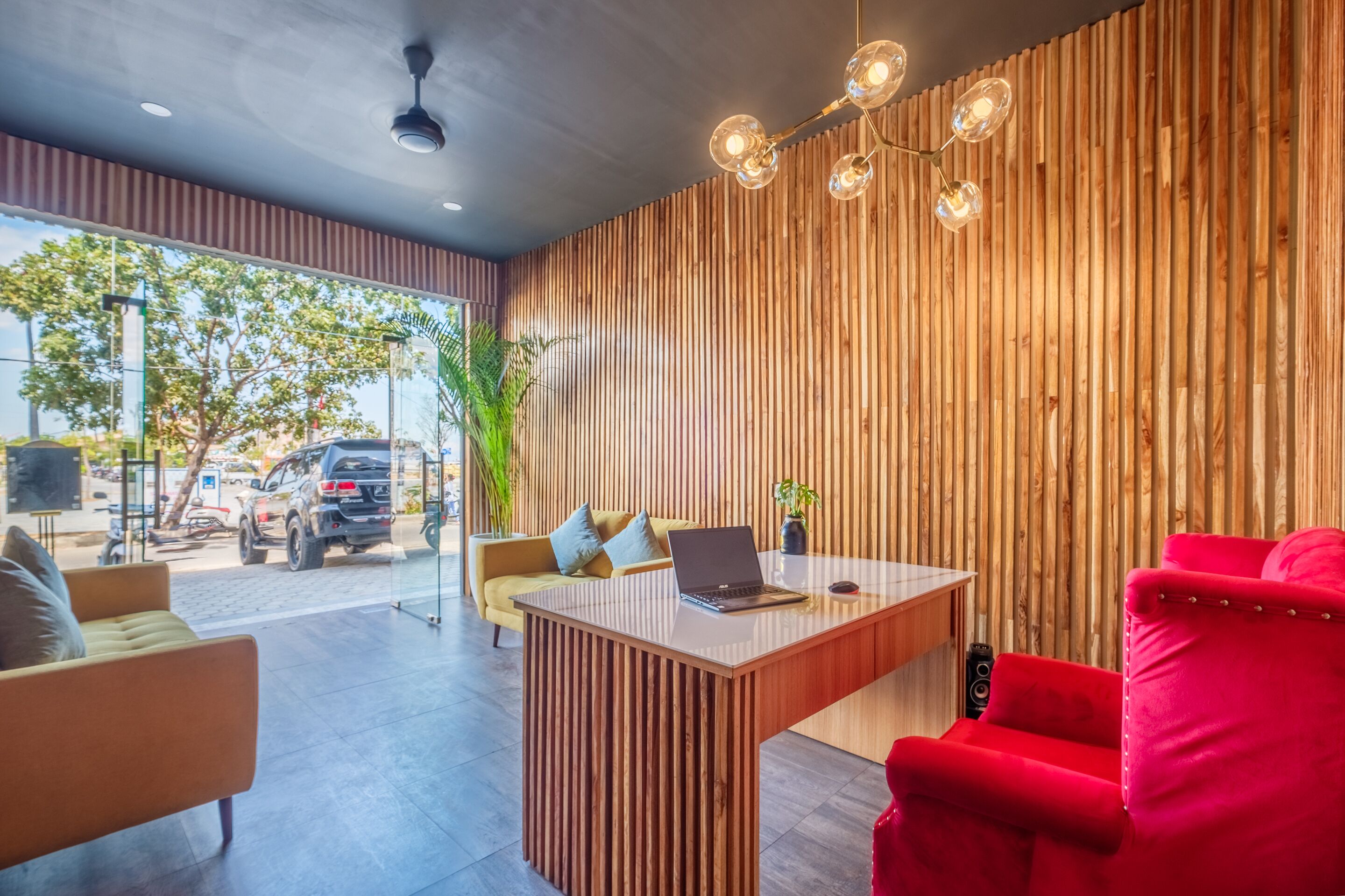 A modern reception area with a wooden slat wall, a desk with a laptop, a red armchair, and a yellow sofa.